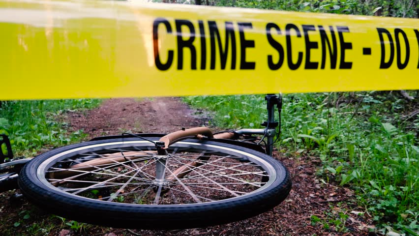 Crime documentary type footage of a bicycle laying on a forest floor with a yellow crime scene tape in front of it while the bikes front wheel is spinning. Bike is on a forest road with no people.