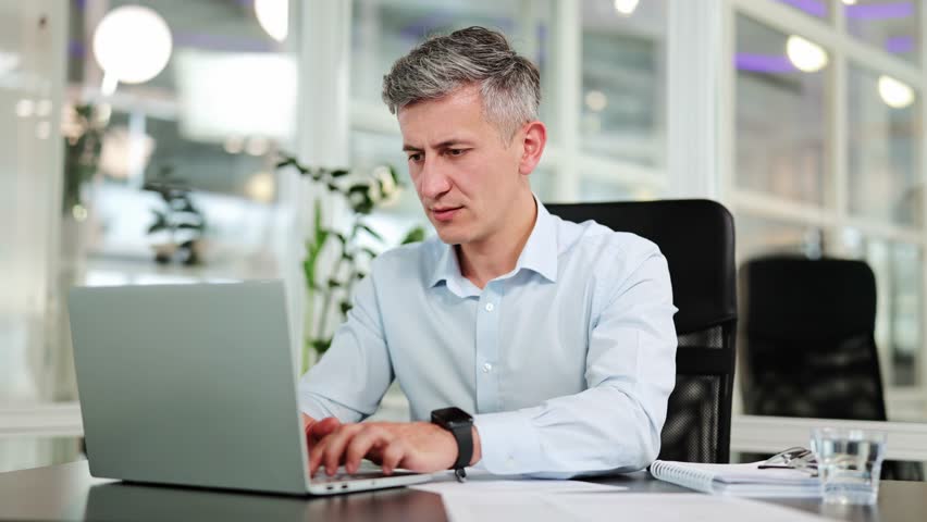 Concerned middle-aged businessman, dressed in light blue button-up shirt, suffers from toothache or jaw pain while working on laptop in bright office, expressing discomfort.
