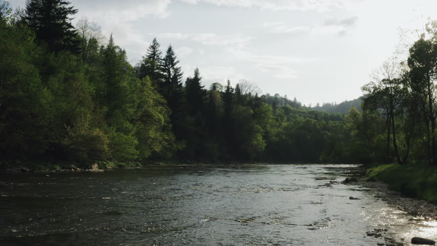 Establishing shot of gently flowing shallow river reflecting warm sunset light, dense autumn forest, hills in distance. Aerial low angle shot, dolly left. Bieszczady, Poland.
