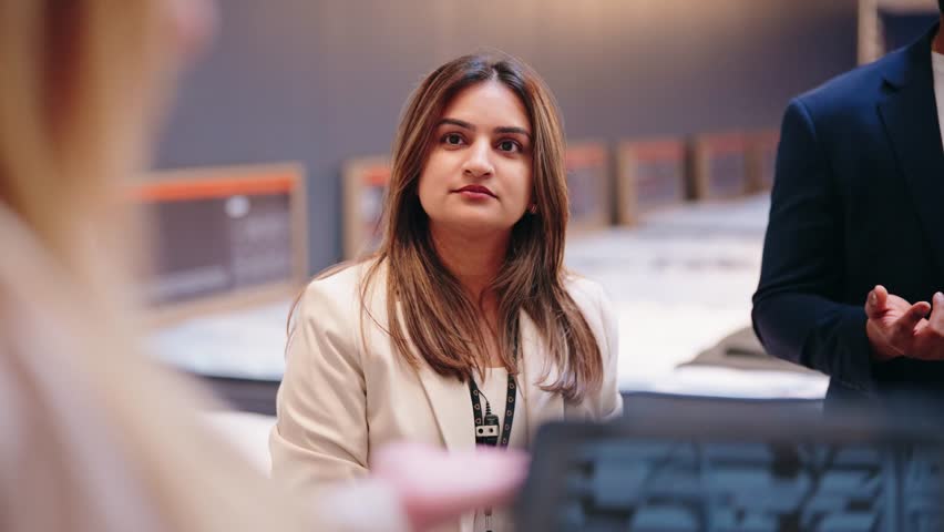 Seated Indian woman nods and laughs with coworker across mattress in store