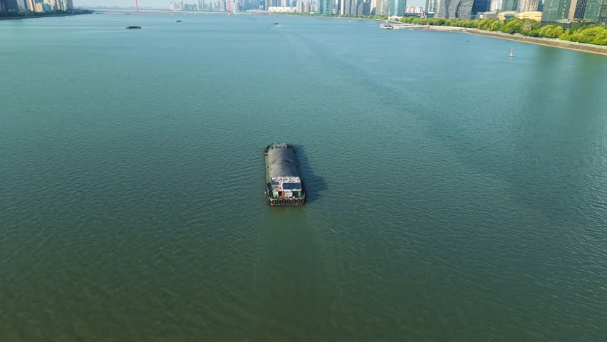 Aerial view of barge navigates river in urban setting with modern skyscrapers during clear day. Hangzhou, China.