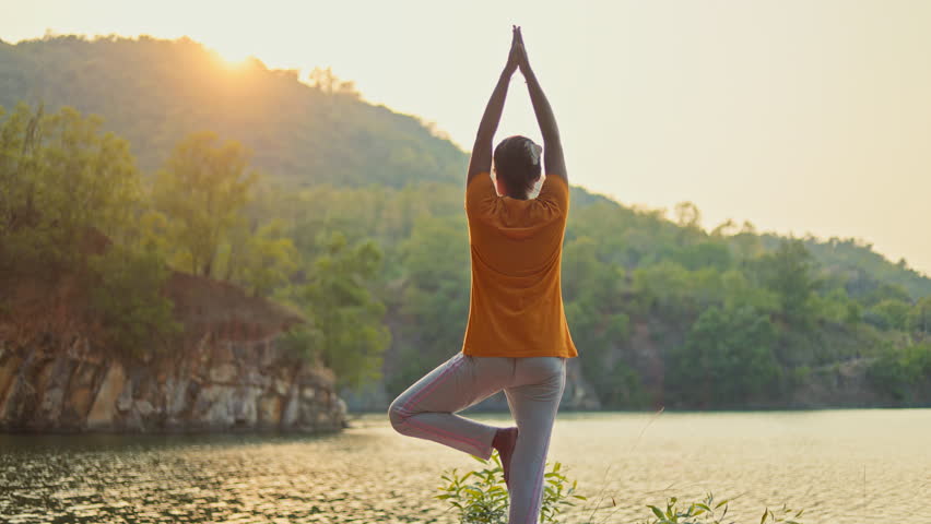 Indian woman practicing vrikshasana near a lake facing towards morning sunlight during golden hour.
