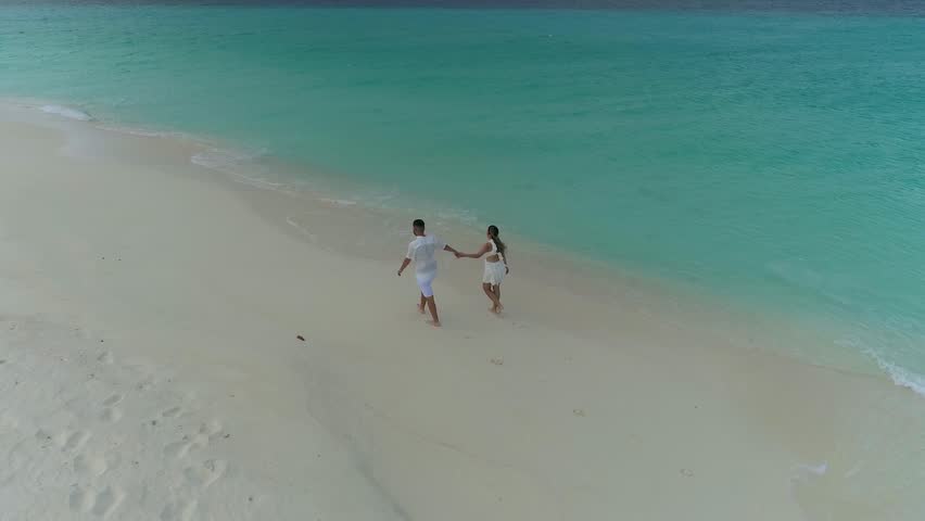 Aerial view of a couple walking on a private Caribbean beach in Los Roques, Venezuela, drawing a heart in the white sand by the turquoise sea.