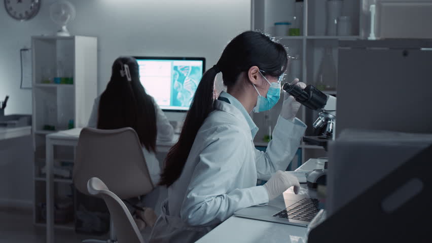 Medium shot with lens flare of focused Asian female scientist in white coat, protective mask, gloves examining cell culture in microscope, typing on laptop, working on university thesis in laboratory - Powered by Shutterstock - Get 15% off with code: PIKWIZARD15