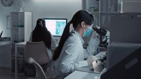 Medium shot with lens flare of focused Asian female scientist in white coat, protective mask, gloves examining cell culture in microscope, typing on laptop, working on university thesis in laboratory - Powered by Shutterstock - Get 15% off with code: PIKWIZARD15