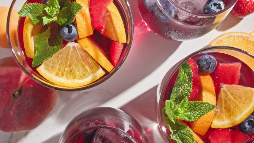 closeup of rotating red sangria glasses with fresh fruit pieces, ice and berries, decorated with mint leaf, top view, on sunny light background