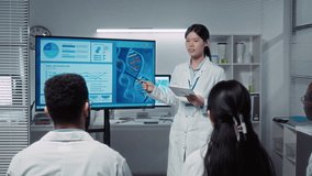 Medium full shot of confident young Asian female student in white coat presenting research paper on genetics, bioengineering and DNA to group of multiethnic colleagues and professor in laboratory - Powered by Shutterstock - Get 15% off with code: PIKWIZARD15