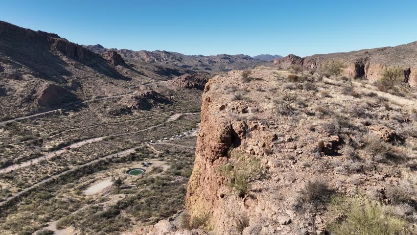 Aerial view of rugged mountains and valley, punctuated by sparse vegetation and a distant lake under a clear sky, Apache Junction, Arizona, United States.