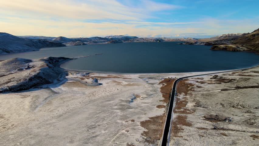 Aerial view of a dark road cuts through a snow-dusted landscape beside a tranquil lake, contrasting with rugged mountains, Grindavíkurbaer, Iceland.