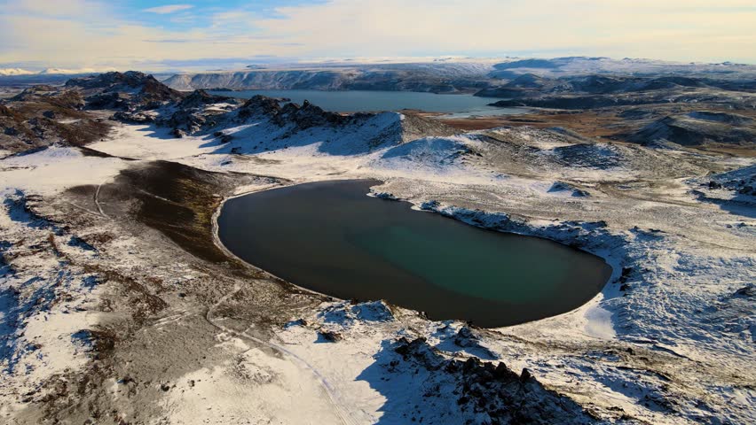 Aerial view of a serene landscape featuring dark lakes surrounded by rugged, snow-dusted terrain under a soft sky, Grindavíkurbaer, Iceland.