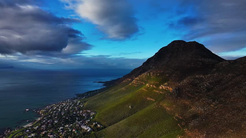 Aerial view of dramatic mountain meeting the coastline, with dark clouds contrasting against the bright blue sky, Cape Town, Western Cape, South Africa.
