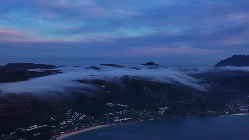 Aerial view of the fog rolling over the mountains and coastline with a blue ocean and sky in the background, Cape Town, Western Cape, South Africa.