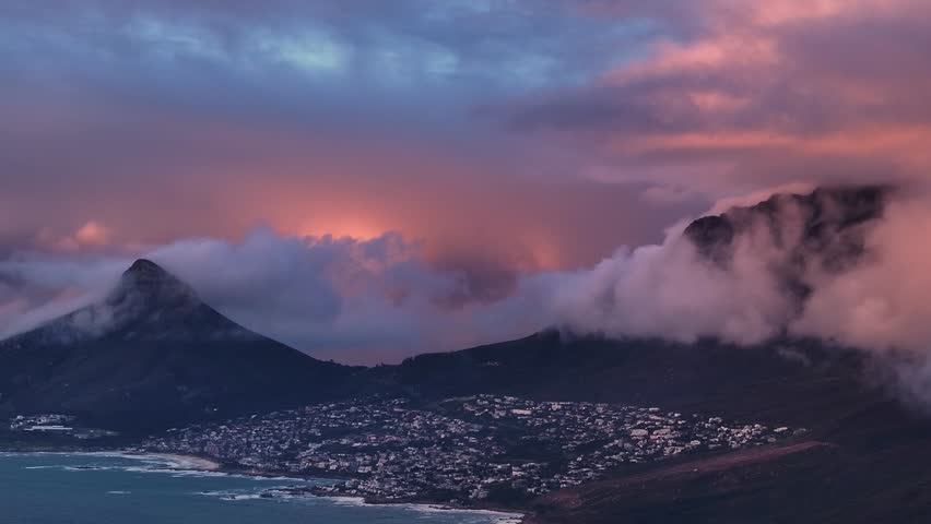 Aerial view of mountains shrouded in ethereal clouds, contrasting with the town below, bathed in the soft glow of twilight, Cape Town, Western Cape, South Africa.