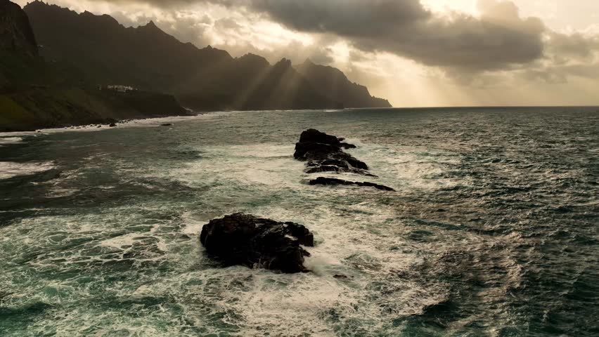 Aerial view of the dramatic coastline with sunlight piercing through the clouds, illuminating the dark, rocky cliffs and choppy ocean waters, Santa Cruz de Tenerife, Spain.
