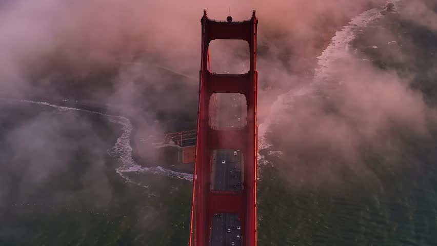 Aerial view of the iconic Golden Gate Bridge shrouded in ethereal fog, its red hue contrasting with the cool ocean and the soft, pink-tinged sky, San Francisco, California, United States.