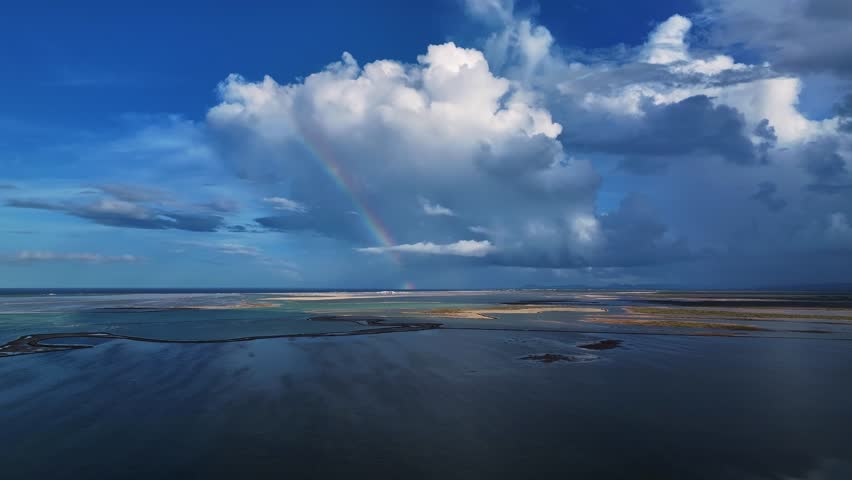 Aerial View of a vibrant rainbow arcing across the sky after a storm, painting vivid colors over the tranquil waters. The clouds are big and fluffy.