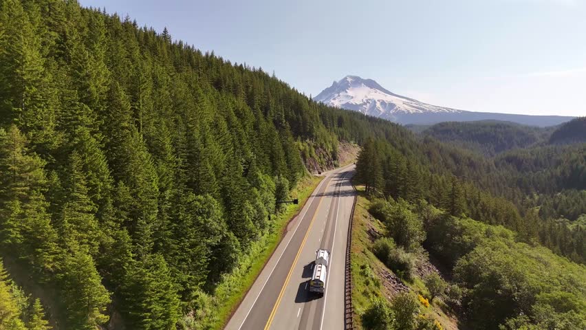 Aerial view of the winding highway cutting through lush green forests, leading to the snow-capped peak of Mount Hood, Government Camp, Oregon, United States.