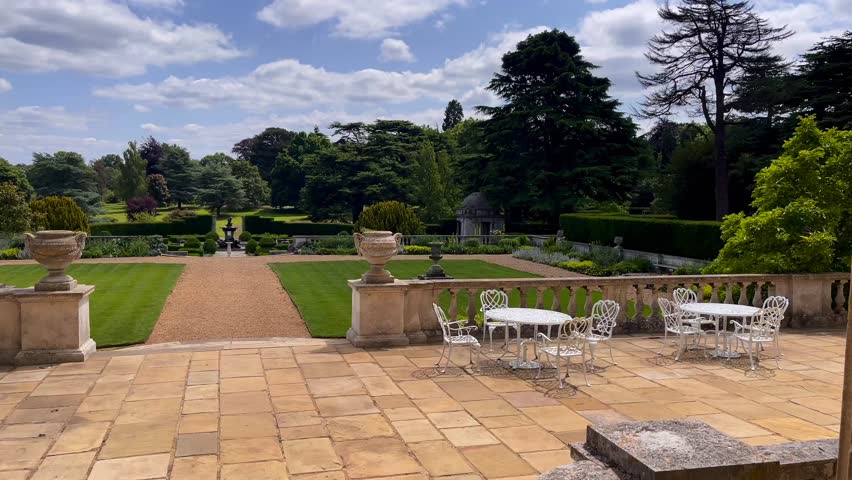 English garden terrace with ornate urns, iron seating and manicured lawns leading into symmetrical formal hedges, blue sky and clouds above, slow motion