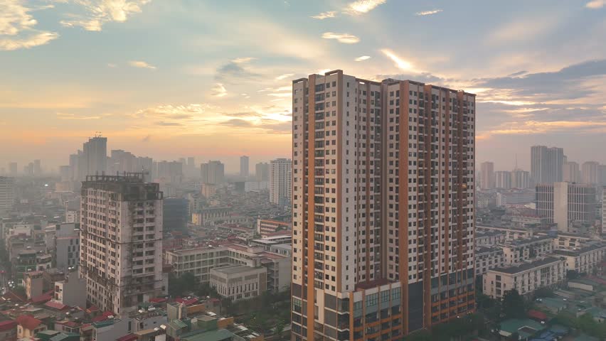 An atmospheric aerial view of Hanoi, Vietnam, at sunset, bathed in the warm glow of the sun through a soft haze. The sprawling urban landscape stretches towards the horizon, with distant lakes or rive