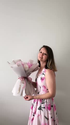 Woman wearing elegant floral dress standing with vibrant flower bouquet, radiating happiness and serenity while gently holding blossoms against pristine white background