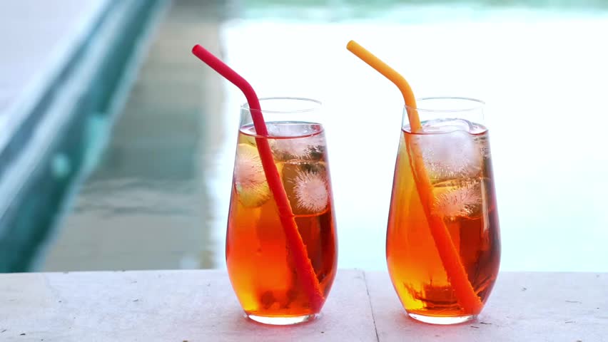Close up horizontal clip of two homemade Aperol spritz cocktails with ice and colorful silicone straws sit on a sunny poolside ledge, perfect for summer vibes. Private swimming pool
