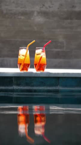 Two homemade Aperol spritz cocktails with ice and colorful silicone straws sit on a sunny poolside ledge, perfect for summer vibes. Vacation on private villa 
