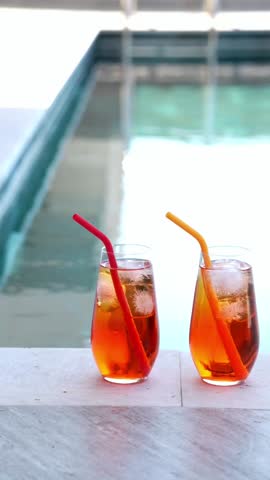 Vertical video of two homemade Aperol spritz cocktails with ice and colorful silicone straws sit on a sunny poolside ledge, perfect for summer vibes. Private swimming pool
