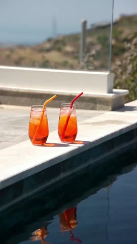 Two homemade Aperol spritz cocktails with ice and colorful silicone straws sit on a sunny poolside ledge, perfect for summer vibes. Vacation on private villa 
