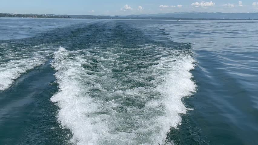 Motorboat leaving foamy wake on peaceful blue lake with distant forested shore and mountains under sunny sky, perfect for travel, adventure, boating, summer leisure, nature and water sports themes