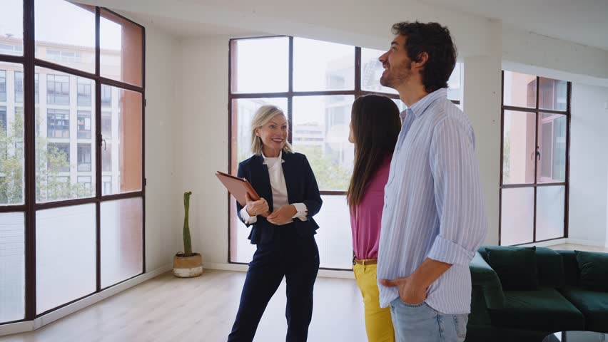 Woman in business attire presents a spacious apartment to a couple, highlighting large windows and urban views, showcasing potential living space