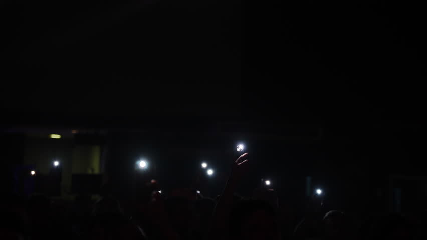 Concert crowd capturing musical moment, smartphone flashlights creating luminous atmosphere with synchronized energy and shared excitement