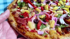 Close up macro dynamic view holding a slice of colorful vibrant pizza with red, green, yellow peppers, mushrooms, vegetables, pineapple, onions, and pepperoni toppings on a picnic table background - Powered by Shutterstock - Get 15% off with code: PIKWIZARD15