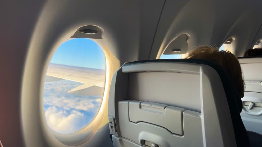 View on the fragment of the airplane cabin with passengers during the flight and a plane porthole through which you can see the wing of the plane and the blue sky with clouds in sunset.