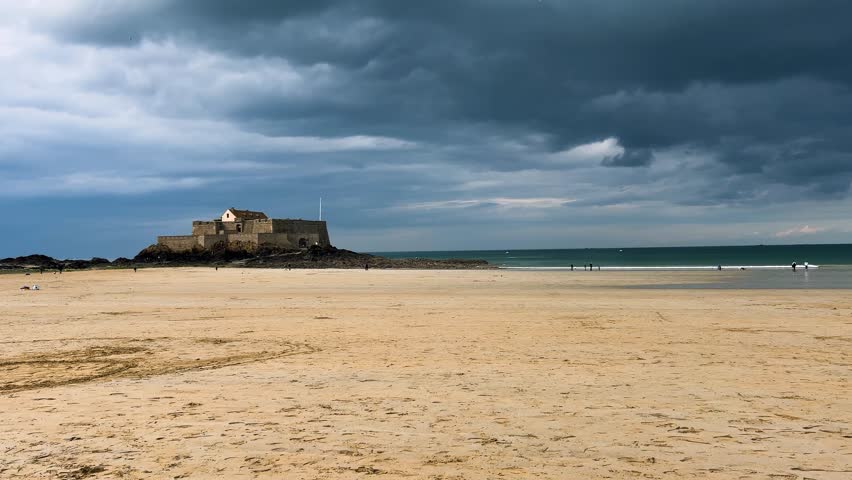 View of the Plage de l’Eventail sandy beach, the English Channel and the National Fort illuminated by the sun under a stormy sky in Saint Malo, Brittany, France.