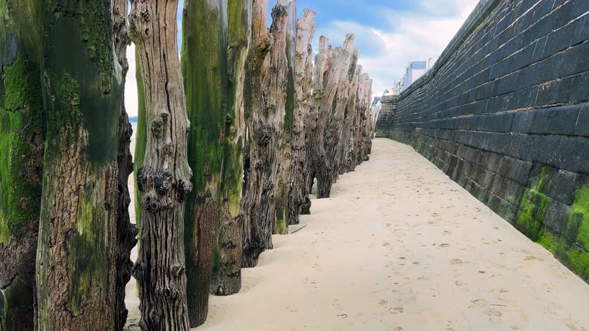 A walk along the Plage de l’Eventail sandy beach past the stone city wall and wooden pilings of the breakwaters overlooking the English Channel in Saint Malo, Brittany, France.