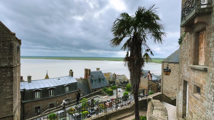 A tall palm tree sways in the strong wind against the backdrop of the English Channel at low tide on a cloudy spring day. Top view on the architecture of Mont-Saint-Michel Abbey, Normandy, France.