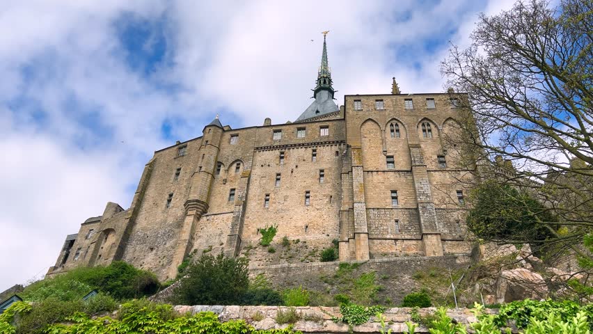 View of the Mont-Saint-Michel Abbey walls and seagulls that soaring in the blue sky with clouds on a windy spring day. Normandy, in the department of Manche, France.