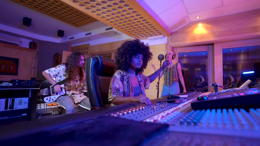 Music producer managing sound levels on audio mixing console while musicians play guitar and sing