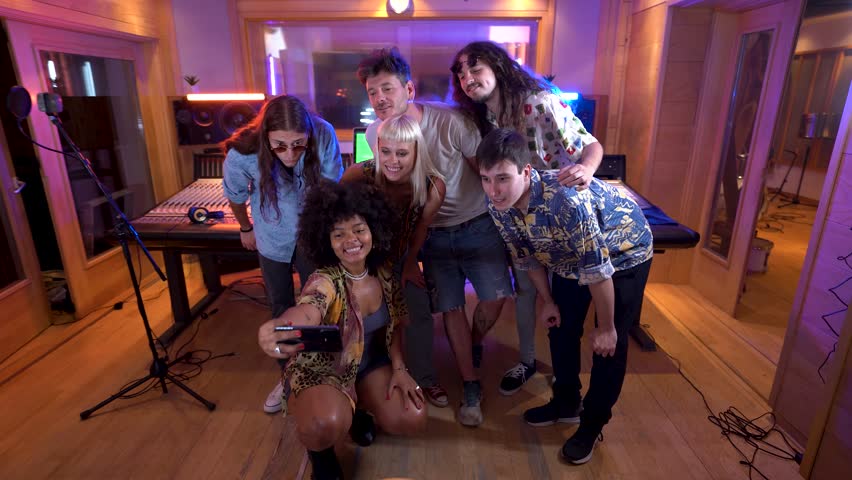 Diverse group enjoying a lively selfie session inside a modern music studio with colorful lights and equipment in the background