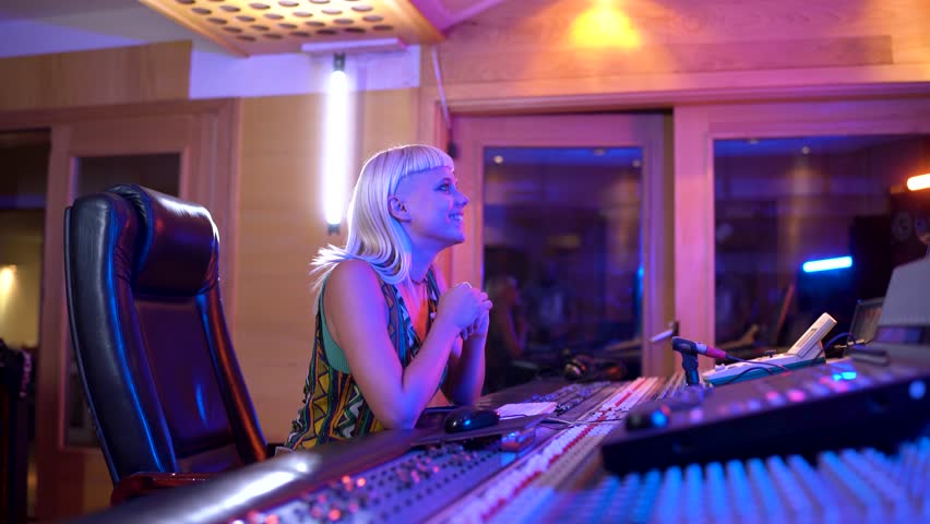 Focused female music producer works diligently in a colorful, illuminated recording studio, adjusting sound equipment and making creative decisions