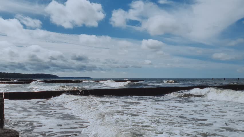 Serene seaside landscape of Baltic sea coast with stormy waves rolling on empty beach and old wooden breakwater under dramatic cloudy sky at summer day. No people seascape natural background.