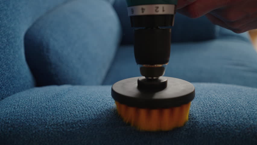 Professional cleaning service worker using cordless screwdriver with round brush attachment to clean blue sofa, providing efficient and thorough upholstery cleaning, close-up. Shooting in slow motion.