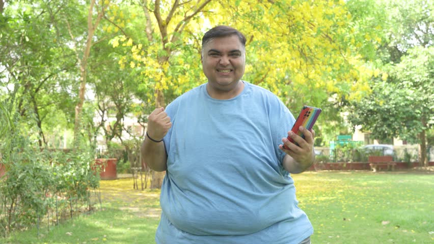 Excited overweight fat indian man win lottery on smart phone at summer park. Plus size obese guy celebrate. Weight loss. Good luck. Wireless technology. 5g