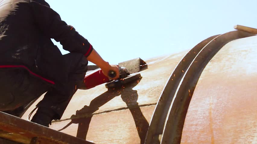 A rear view shows a man hitting a large metal tank with a hammer. A metal part breaks off on impact. Filmed in hot weather and strong sunlight. Full HD video.