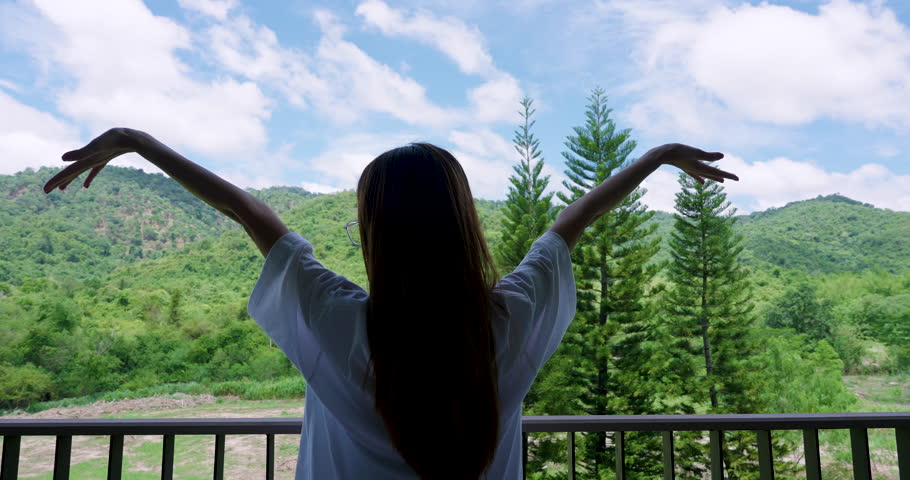 Asian adult woman stretches arms on balcony in slowmotion facing mountain view during peaceful morning at home showing soft routine healthy mindset and calm relaxed lifestyle outdoors