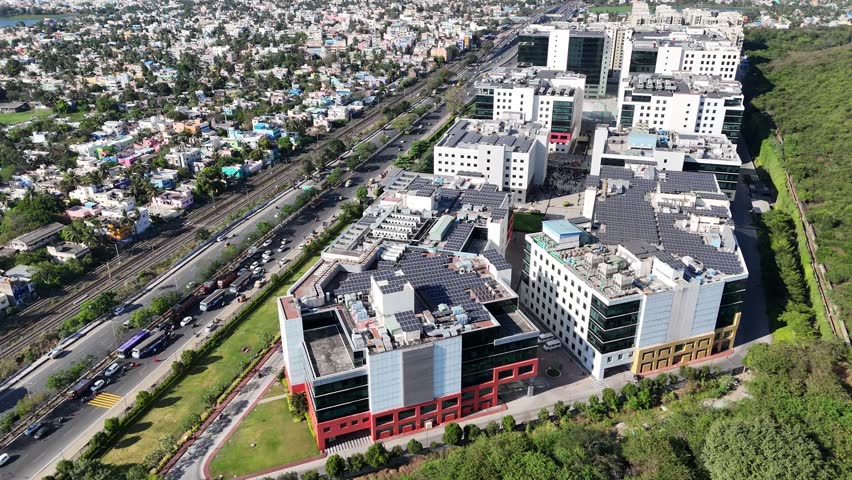 Aerial view of a modern Chennai business park with solar-panelled buildings. It features busy roads, railway, and residential areas on one side, with green hills on the other, shows urban development.