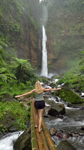 Blonde woman walks on bamboo bridge toward the majestic Kapas Biru Waterfall in East Java, Indonesia, surrounded by lush jungle, mossy rocks, and flowing river.