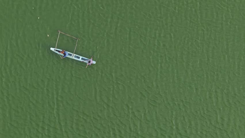 Aerial top-down view of two fishermen paddling a traditional boat in vibrant green water.