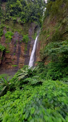 Cinematic FPV drone shot diving through scenic Kapas Biru waterfall cascading down a tall cliff into lush green canyon, surrounded by tropical rainforest in East Java, Indonesia.