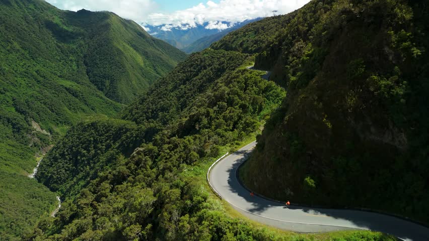Group of adventurous tourists cycling downhill on a winding mountain road through a lush green valley as part of the Inca Jungle Tour in Peru - A stunning slow parallax drone shot